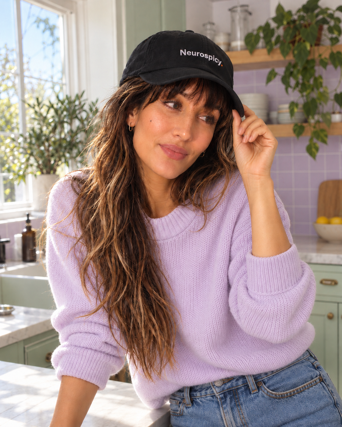 Woman in a lilac jumper wearing the black Neurospicy. embroidered cap in a pastel kitchen